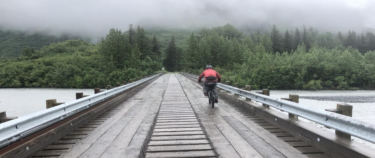 A bikepacker rides along the Alaskan Pipeline access road in misty weather, highlighting key bikepacking pre-trip considerations for remote and unpredictable conditions.